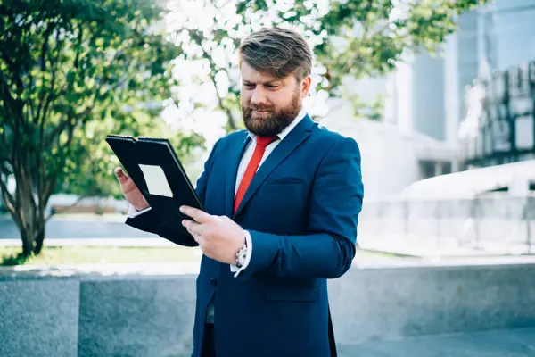 Concern serious doubtful businessman in suit looking at paperwork in folder not happy with work results standing on street near business center