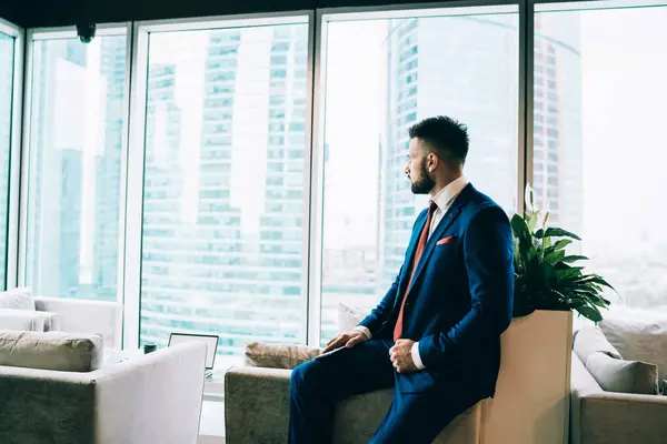 Side view of contemporary executive man in dark blue suit and red tie sitting on chair in modern office center lounge and looking thoughtfully at window