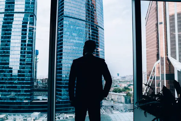 Modern ofisin panoramik penceresinden şehir merkezine bakarken başarılı, düşünceli iş adamının siluetinin arka planda kalan görüntüsü.