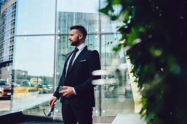 Takım elbiseli, akıllı telefon ve güneş gözlüklü adam ofis binasının önünde dikilirken yüzünü çeviriyor.