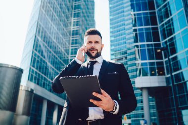 Gündüz güneşli bir günde gökdelenlerin dışında akıllı telefon görüşmeleri yaparken planlayıcısı gülümseyen ve kameraya bakan kendine güvenen bir iş adamı.