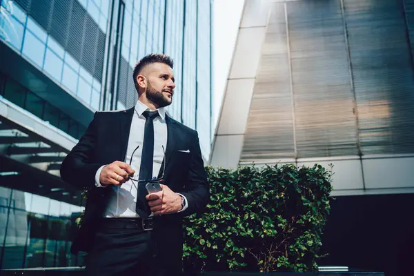 Güneş gözlüklü ve akıllı telefonlu zarif siyah takım elbiseli neşeli, kendine güvenen işadamının şehir merkezindeki modern yüksek katlı ofis binalarına karşı caddelerde durup gelecek için stratejisini geliştirirken...