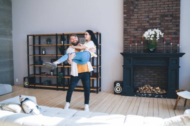 Genç ve mutlu bir çift taşındıktan sonra yeni dairenin ferah oturma odasında ısınırken koca karısını kucağında taşıyor.