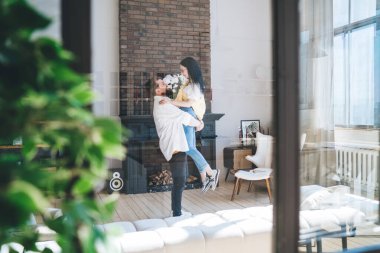 Gündelik giysiler içinde gülümseyen, kucaklaşan ve birbirine bakan insanların geniş modern oturma odasında durduğu bir pencereden yan bakışı.