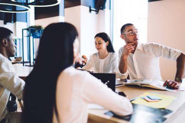 Yuvarlak gözlükleri ve kol saati olan genç yaratıcı adam dikkatle başka tarafa bakıyor ve modern ofisteki çeşitli iş gruplarıyla fikirleri tartışıyor.