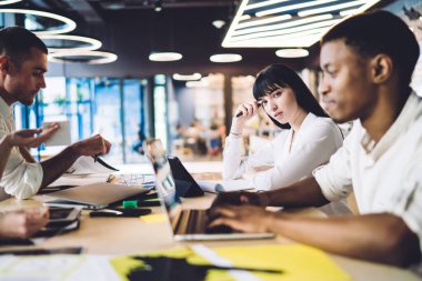 Güzel genç bayan kameraya bakıyor ve ofiste çok ırklı iş arkadaşlarıyla yeni bir proje üzerinde çalışırken oturuyor.