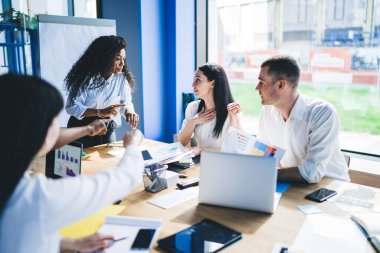 Resmi takım elbiseli konsantre siyahi kadın iş arkadaşlarıyla toplantı sırasında masanın yanında çeşitli aletlerle konuşurken konuşuyor.