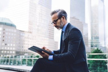 New York binalarının arka planında otururken tablet kullanarak bacak bacak bacak üstüne atmış takım elbiseli ve gözlüklü yetişkin bir adamın yan görüntüsü