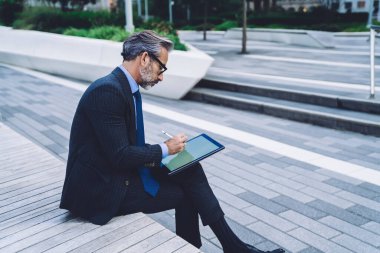 New York sokaklarında bankta otururken Stylus 'la tablet üzerinde çalışırken bacak bacak bacak üstüne atmış orta yaşlı işadamının yan görüntüsü.