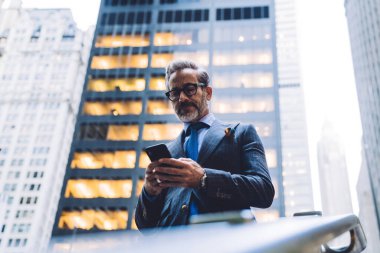 Gözlüklü ve koyu renk takım elbiseli orta yaşlı bir işadamının New York 'taki gökdelenin arka planında telefonla yazı yazması.