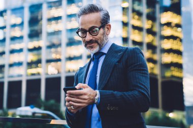 Yakışıklı, gri saçlı, modaya uygun saç kesimli, siyah ceketli, mavi kravatlı ve New York 'ta bulanık arka planda cam çitle akıllı telefondan mesaj atan bir iş adamı.