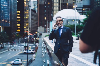 Gözlüklü ve takım elbiseli olgun bir erkek telefonda konuşurken cam çitin yanında New York caddesinin arka planında duruyor.