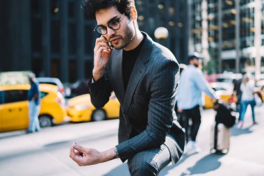 Zarif takım elbiseli ve gözlüklü şık genç adam modern şehrin sokaklarında cep telefonuyla konuşuyor.