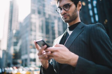 Zarif giyimli, gözlüklü, modern şehrin sokaklarında telefon izleyen, sosyal medyada gezinen modaya uygun bir adamdan.