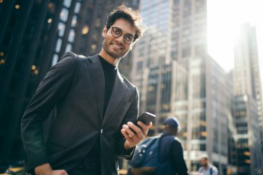 Takım elbiseli ve gözlüklü genç girişimci iş bölgesinde duruyor ve elinde cep telefonuyla kameraya bakıyor.