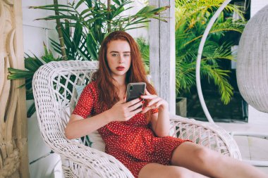 Akıllı telefonlu, kırmızı uzun saçlı, benekli elbiseli, terastaki beyaz koltukta oturan ve kameraya bakan sakin bir genç kadın.