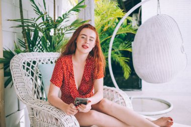 Çekici, kızıl saçlı, kırmızı benekli elbiseli, koltukta oturan, elinde cep telefonu olan ve kameraya bakan bir kadın.