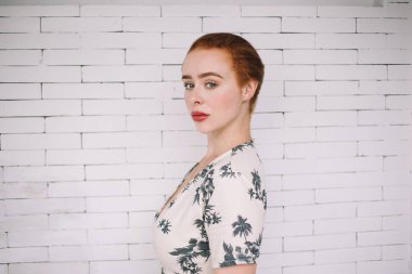 Çiçekli elbiseli, beyaz tuğla duvar arkasında kameraya bakan muhteşem kızıl saçlı kadının yan görüntüsü.