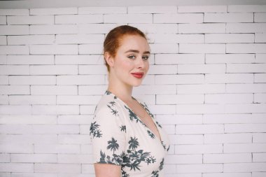 Kameraya gülümseyen beyaz tuğla duvarın arkasında duran, sade elbiseli neşeli, kızıl saçlı kadının yan görüntüsü.