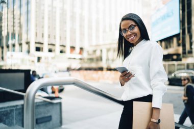 Modern şehrin caddesinde kameraya bakarak akıllı telefon mesajları kullanarak resmi kıyafet ve gözlük takan neşeli Afro-Amerikan kadın.
