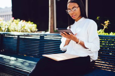 Resmi kıyafetler ve gözlük takan, tablet okuyan genç siyahi bir kadın modern şehrin sokaklarında bankta oturuyor.