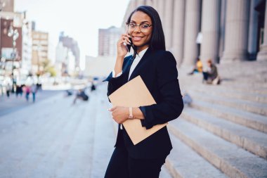 New York caddesinde cep telefonuyla konuşurken elinde randevu defteriyle gülümseyen ve kameraya bakan şık gözlüklü genç Afrikalı Amerikalı kız öğrenci. 