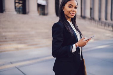 Resmi takım elbiseli neşeli, zarif Afro-Amerikan bayan sosyal medyayı kontrol ediyor. New York sokaklarında kameraya bakıyor.