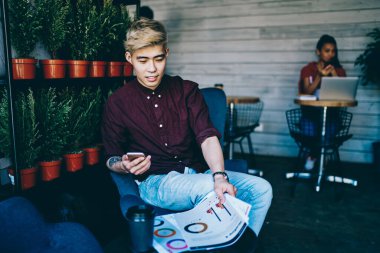Sarışın, eflatun gömlekli Asyalı bir erkek cep telefonuyla çalışma odasında iş arkadaşlarıyla oturmuş şemaları kontrol ediyor.