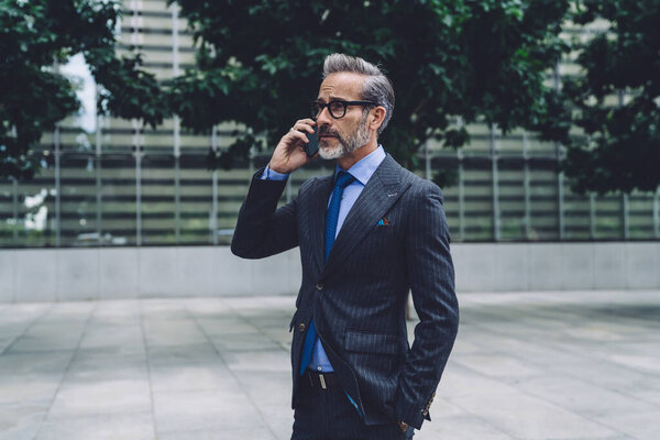 Middle-aged grey-haired busy serious man in official suit and glasses communicating and talking by modern digital smartphone while looking away on urban background