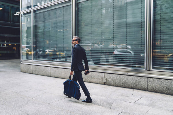 Side view of successful middle aged man in stylish dark business suit walking down city street with big dark blue bag in New York