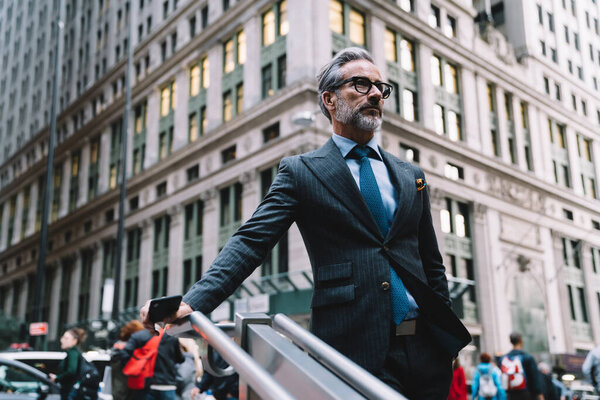 Middle aged man wearing elegant suit and glasses holding mobile phone standing on stairs in downtown area of New York