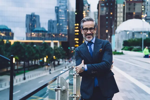 Handsome grinning mature man in glasses and business suit with arms crossed standing against evening New York street while looking at camera