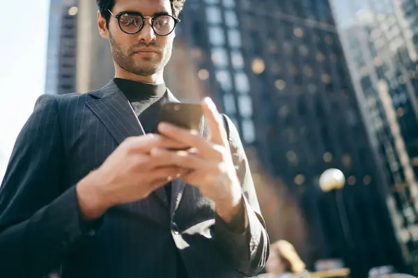 From below of stylish young man in elegant suit and glasses surfing social media watching cellphone in street of modern city