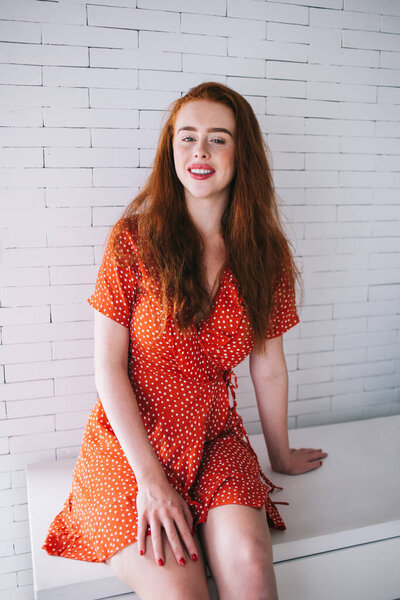 Joyful positive red haired woman in red polka dot dress sitting near white brick wall in bright room and looking at camera