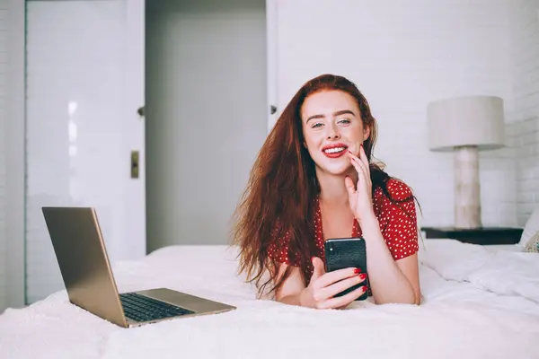 Content satisfied red-haired woman in red dress recreating on soft bed while using laptop and texting on smartphone in trendy contemporary apartment and looking at camera