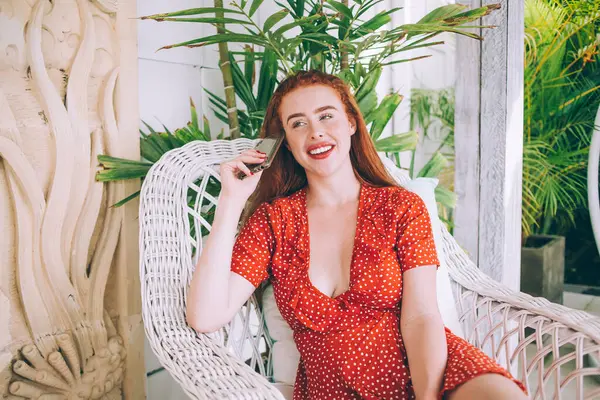 Bright happy woman with red hair and red lips wearing colorful dress with polka dots and chilling on terrace with phone sitting in armchair