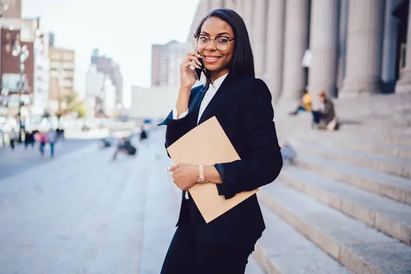 Young African American female student in stylish formal wear and eyeglasses smiling and looking at camera with appointment book in hand while having conversation on mobile phone in New York street