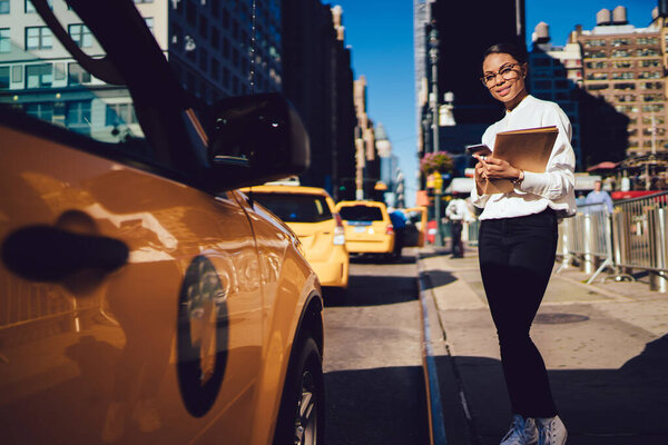 low angle of pleased African American well dressed female student in formal clothing and eyeglasses standing by side of road and looking at camera at modern New York city street 