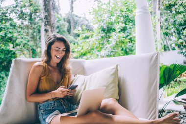 Güzel, gülen esmer kadın. Günlük giysiler içinde. Telefonla mesajlaşırken bahçede dizüstü bilgisayarlı beyaz koltukta oturuyor.