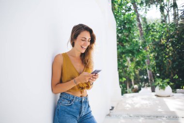 Kıvırcık saçlı, geniş gülümsemeli, cep telefonuyla sörf yapan yeşil ışık bahçesindeki beyaz duvara yaslanan bir kadın.