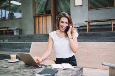Genç neşeli esmer, günlük giysiler içinde açık hava kafeteryasının masasında oturmuş laptopla çalışırken akıllı telefonla konuşuyor ve gülümsüyor.
