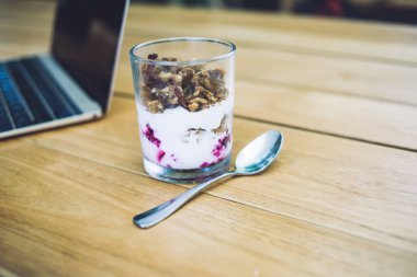 Şeffaf bardakta kuru üzüm ve yoğurt ile iştah açıcı bir tatlı, gümüş çay kaşığı ahşap masaya dizilmiş, boş ekranlı dizüstü bilgisayar.