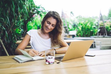 Defterde kayıt yapan meşgul bir kadın dışarıda dinlenirken dizüstü bilgisayar kullanıyor ve bulanık arka planda lezzetli tatlıların tadını çıkarıyor.