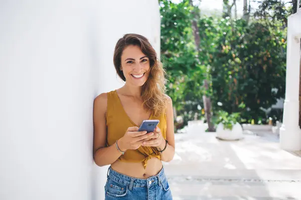 Relaxed pretty woman in crop top leaning against white wall and smiling while surfing on smartphone and looking at camera on blurred background with garden