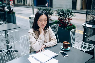 Gündüz vakti bir fincan kahve ve akıllı telefonla sokak terasında oturmuş kitap okuyan modaya uygun genç Asyalı kadınlar.