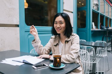Güzel kısa saçlı Asyalı kız öğrenci bej yağmurluk giymiş sokak kafesinde oturmuş elinde kalemle ödev yapıyor kameraya gülümseyen bir kahve içiyor. 