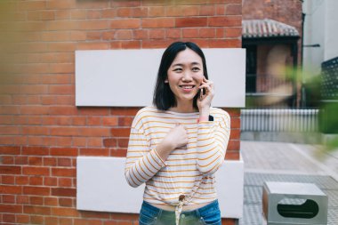 Memnuniyet veren Asyalı kadın, gülümsüyor göğsüne dokunuyor ve kafasını çeviriyor. Şehir sokağındaki tuğla evin köşesinde akıllı telefon konuşmaları yapıyor.