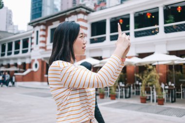 Şehirdeki gezi sırasında meydanın ortasında durup akıllı telefonuyla fotoğraf çeken şaşkın Asyalı kadının yan görüntüsü