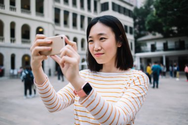 Turistlerle dolu şehir meydanında uzun kollu ve selfie çeken turuncu çizgili tişörtlü sakin Asyalı bir kadın işe alındı.