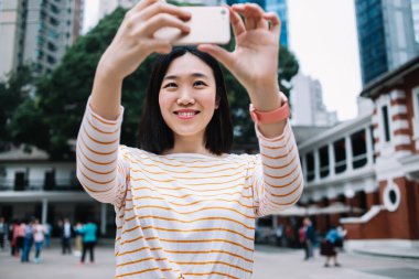 Zeki saati ve kısa siyah saçı olan çekici genç bir kadın şehrin caddesinde dikiliyor ve akıllı telefonuyla gülümserken fotoğraf çekiyor.
